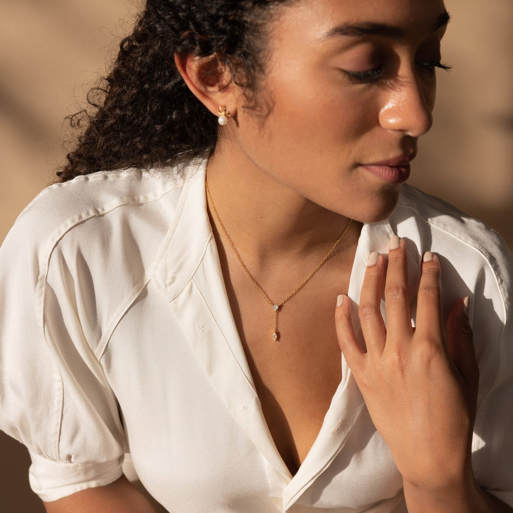 A woman with curly hair in a white shirt wears the Leena Diamond Lariat Necklace, a gold chain, as she looks down with her hand on her chest.