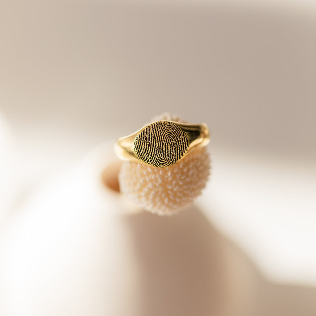The Fingerprint Signet Ring, featuring a gold finish, is displayed on a textured white surface under soft natural light—an elegant piece of memorial jewelry.