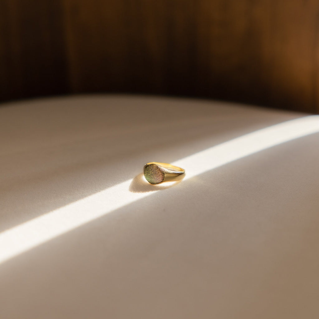 The Fingerprint Signet Ring in gold rests on a sunlit surface, its personalized design creating a memorable keepsake and a unique remembrance gift against the shadowed background.