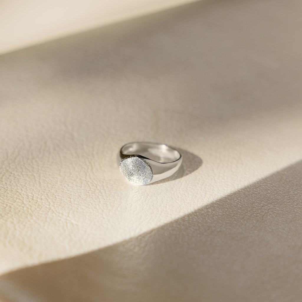 The Fingerprint Signet Ring, finished in silver, sits on a textured light beige surface bathed in soft sunlight—an elegant choice for personalized memorial jewelry.