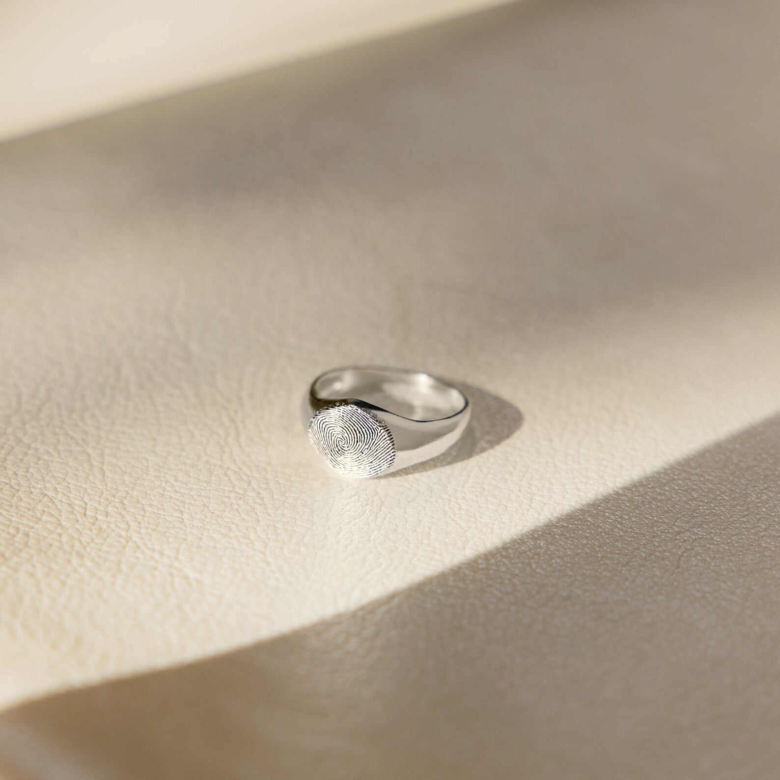 The Fingerprint Signet Ring, finished in silver, sits on a textured light beige surface bathed in soft sunlight—an elegant choice for personalized memorial jewelry.