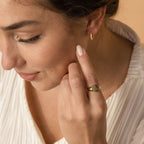 A woman with light nail polish touches her face, wearing a Fingerprint Signet Ring and a small gold hoop earring—a unique piece of memorial jewelry.