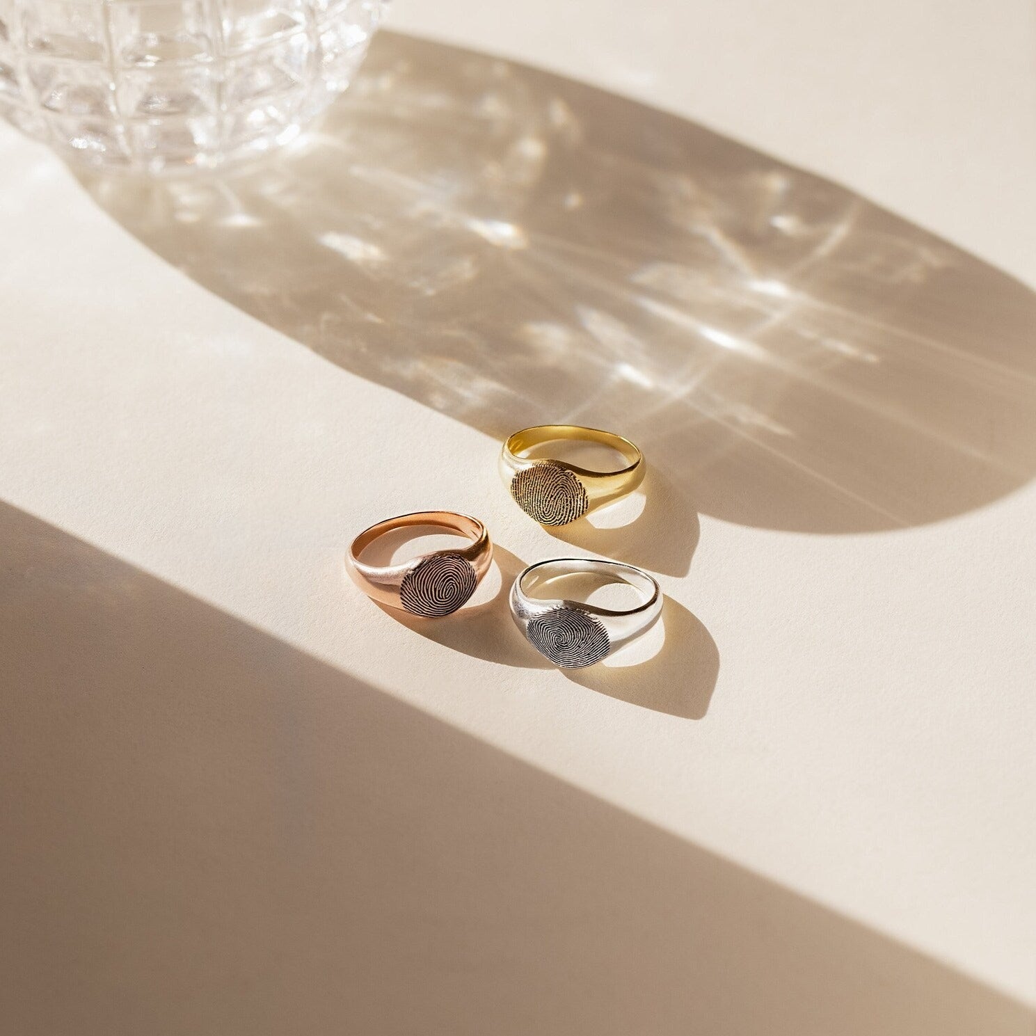 The Fingerprint Signet Ring is available in gold, rose gold, and silver, displayed on a beige surface with sunlight and glass reflections—ideal as a remembrance gift or as personalized memorial jewelry.