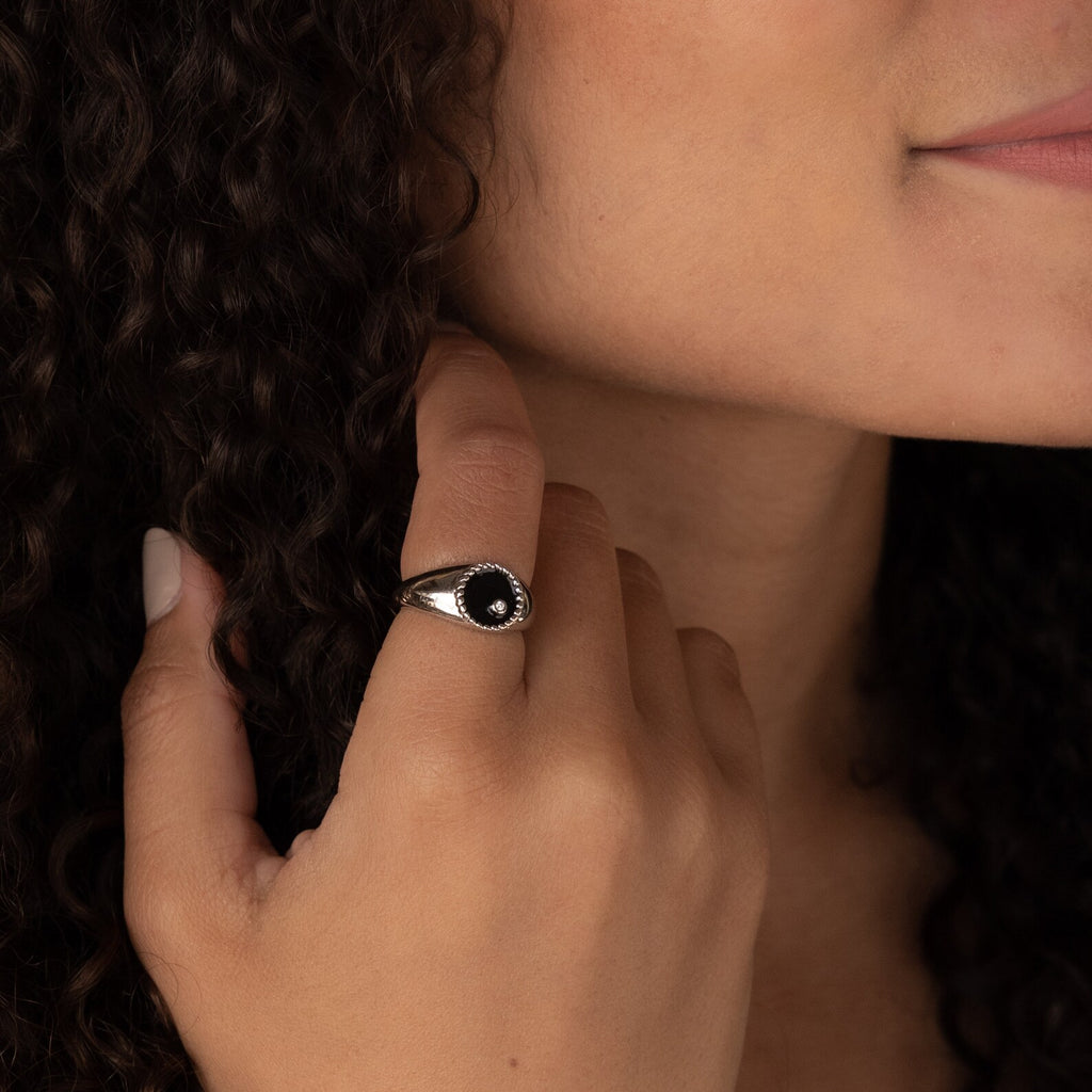 A woman with curly hair wearing the Natasha Black Signet Ring gently touches her hair and smiles slightly.