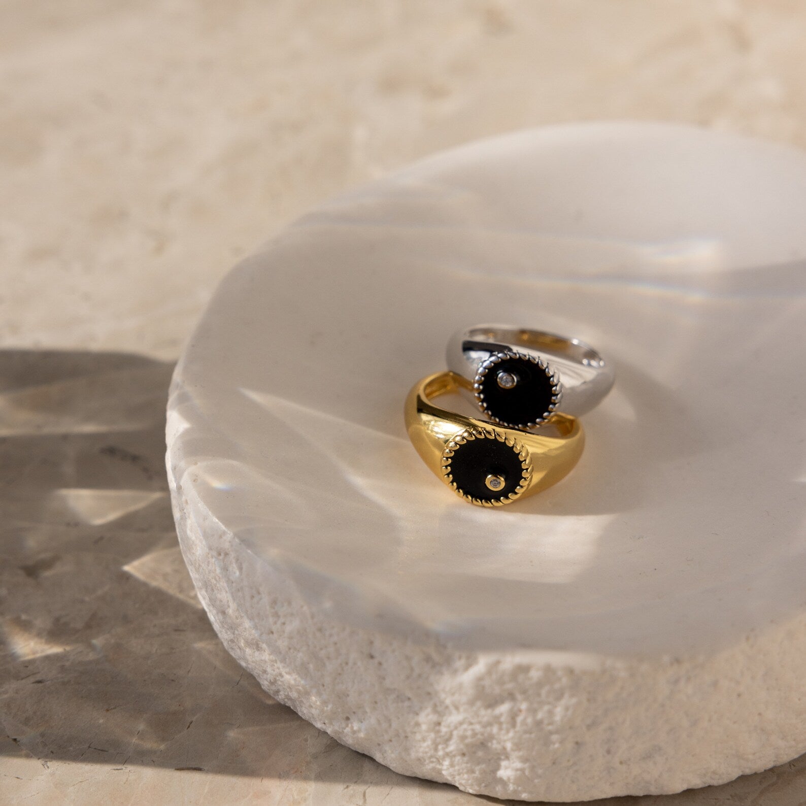 Two Natasha Black Signet Rings in gold and silver, each featuring black stone accents, are displayed on a textured white dish.