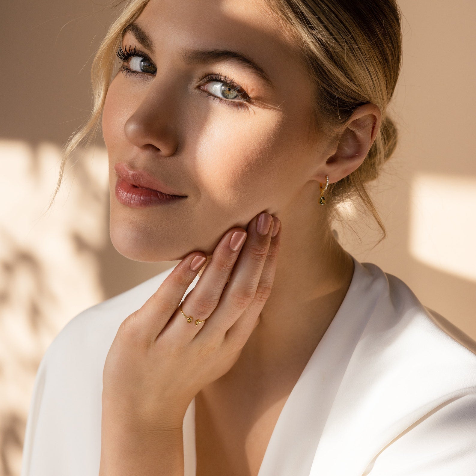 A woman with blonde hair in a white top gently touches her face, softly lit and wearing gold jewelry, including the Duo Marquise Baguette Ring.