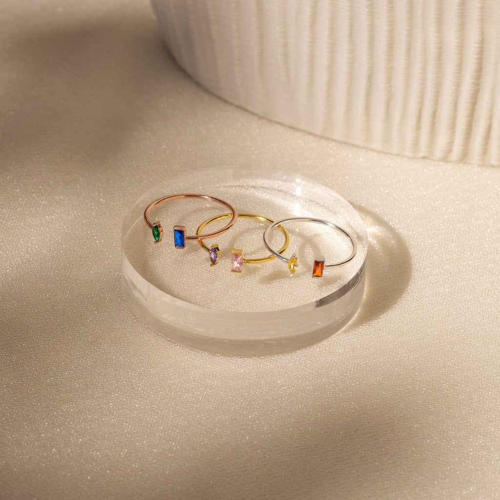 The Duo Marquise Baguette Ring features delicate gold bands adorned with colorful rectangular gemstones, elegantly displayed on a clear round platform in soft light.