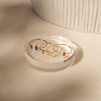 The Duo Marquise Baguette Ring features delicate gold bands adorned with colorful rectangular gemstones, elegantly displayed on a clear round platform in soft light.