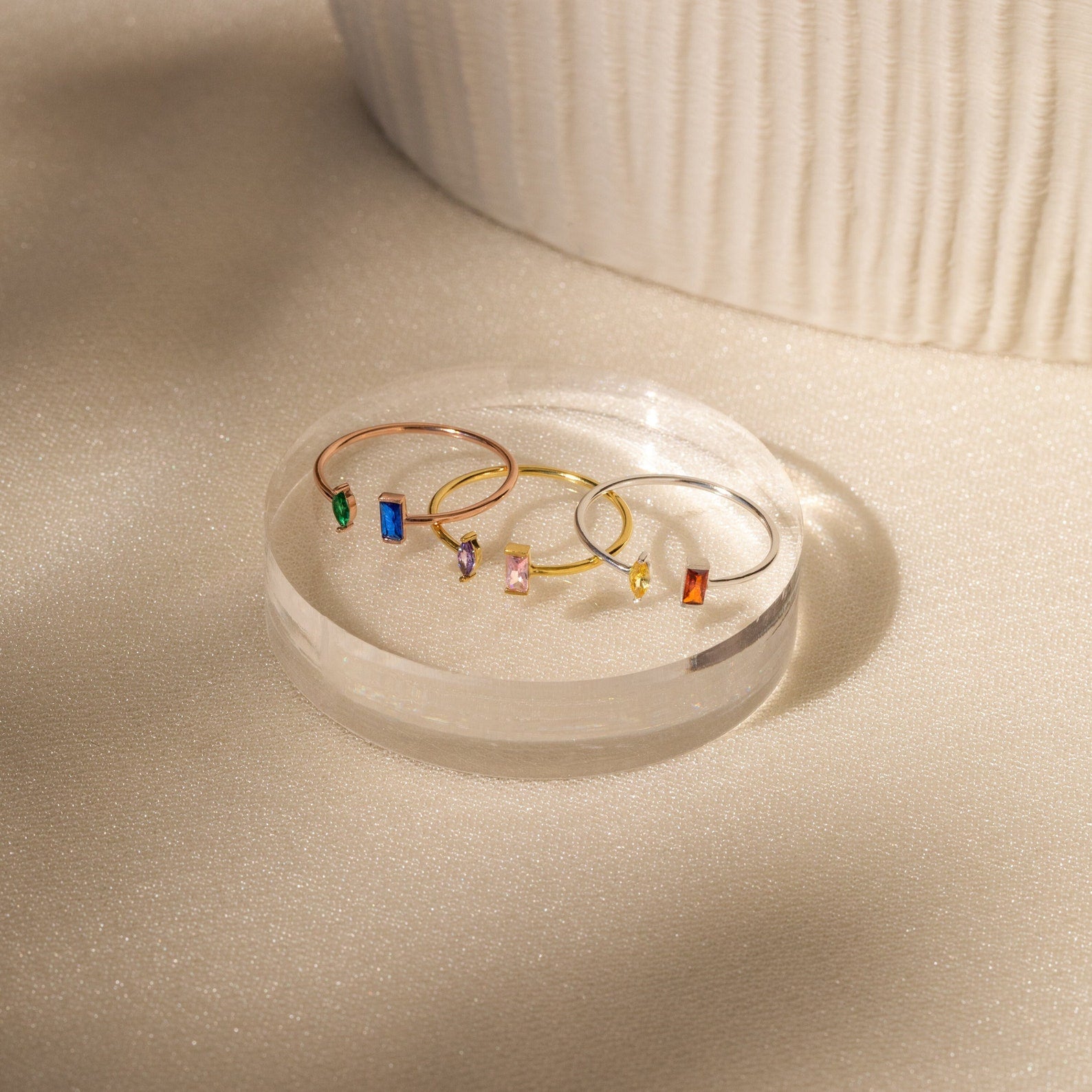 The Duo Marquise Baguette Ring features delicate gold bands adorned with colorful rectangular gemstones, elegantly displayed on a clear round platform in soft light.