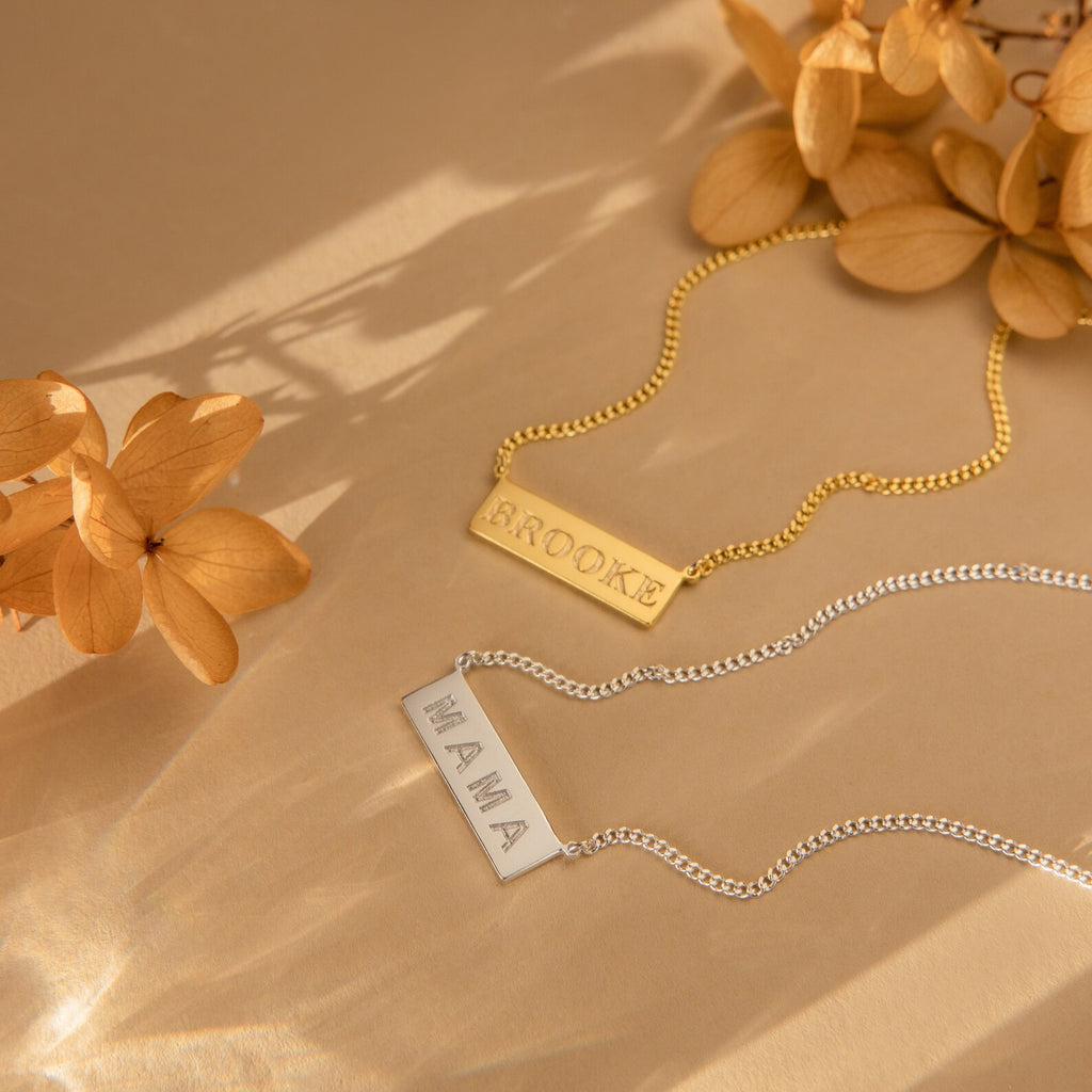 Two Dainty Bar Name Necklaces—one gold with BROOKE and one silver with MAMA—are displayed beside dried flowers, perfect for a personalized touch.