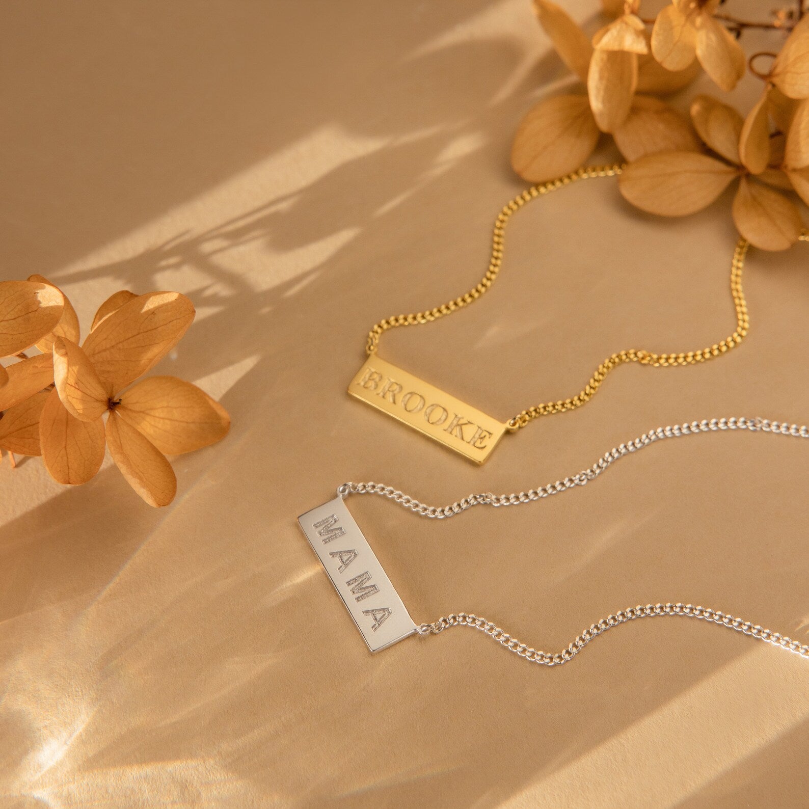 Two Dainty Bar Name Necklaces—one gold with BROOKE and one silver with MAMA—are displayed beside dried flowers, perfect for a personalized touch.