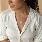A woman in a white blouse wears the Eliza Oval Locket Necklace, sitting and gazing to the side.