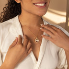 Smiling woman in a white blouse touches a gold Crystal Locket Necklace with a round pendant.