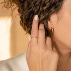 A woman with curly hair wears gold hoop earrings and the Duo Marquise Birthstone Ring, touching her face with manicured fingers.