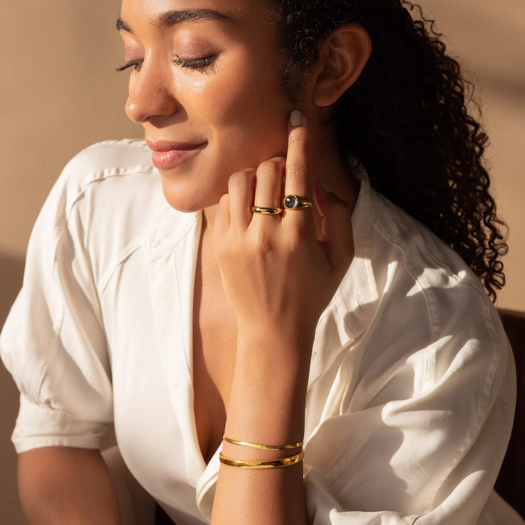A woman in a white blouse wearing gold jewelry, including the Alyssa Cuff Bracelet, closes her eyes and touches her face under warm lighting.