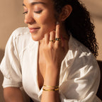 A woman in a white blouse wearing gold jewelry, including the Alyssa Cuff Bracelet, closes her eyes and touches her face under warm lighting.