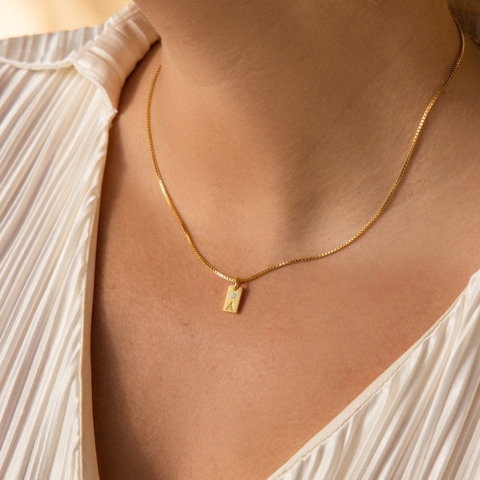 Close-up of a woman wearing the Mini Birthstone Tag Necklace, featuring a gold engraved rectangular pendant, paired with a white pleated blouse.