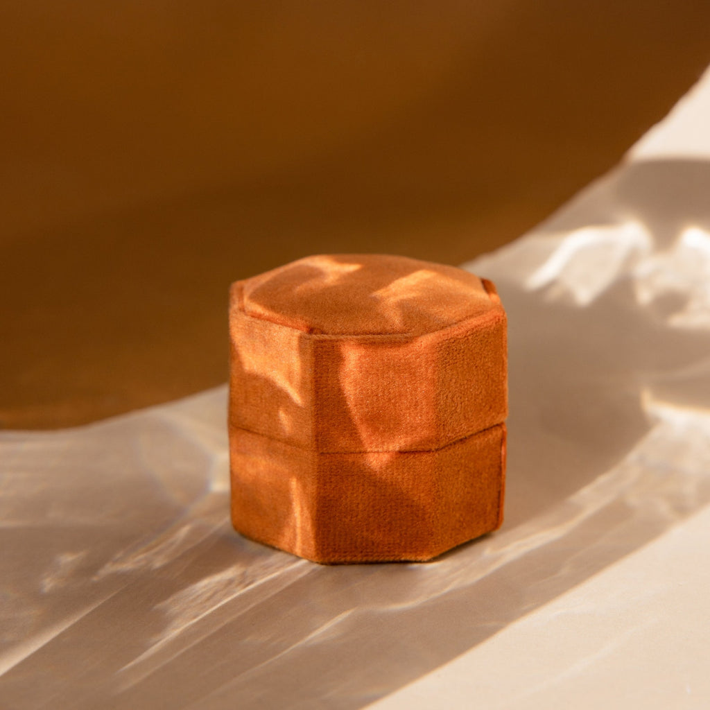 The 3 Slot Velvet Ring Box features a hexagonal, rust-colored design, displayed on a soft, light surface with warm natural lighting and gentle shadows.