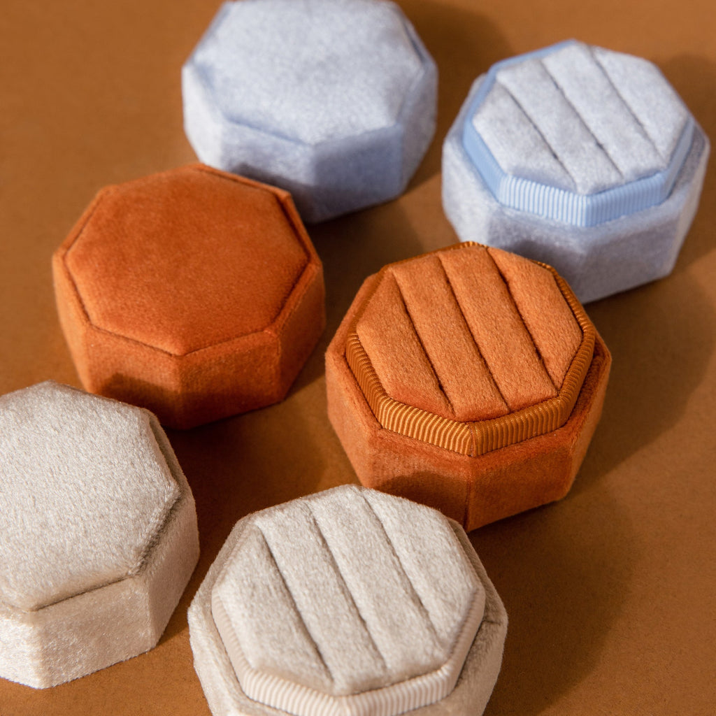 The 3 Slot Velvet Ring Boxes in hexagonal and octagonal shapes, available in beige, orange, and light blue, are arranged on a brown surface for a stylish jewelry organizer display.