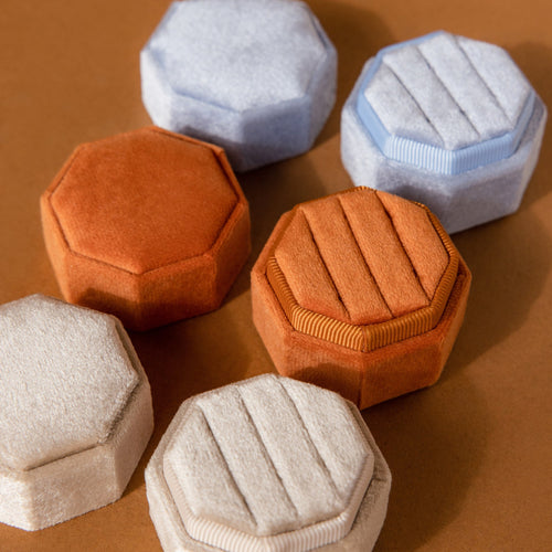 The 3 Slot Velvet Ring Boxes in hexagonal and octagonal shapes, available in beige, orange, and light blue, are arranged on a brown surface for a stylish jewelry organizer display.