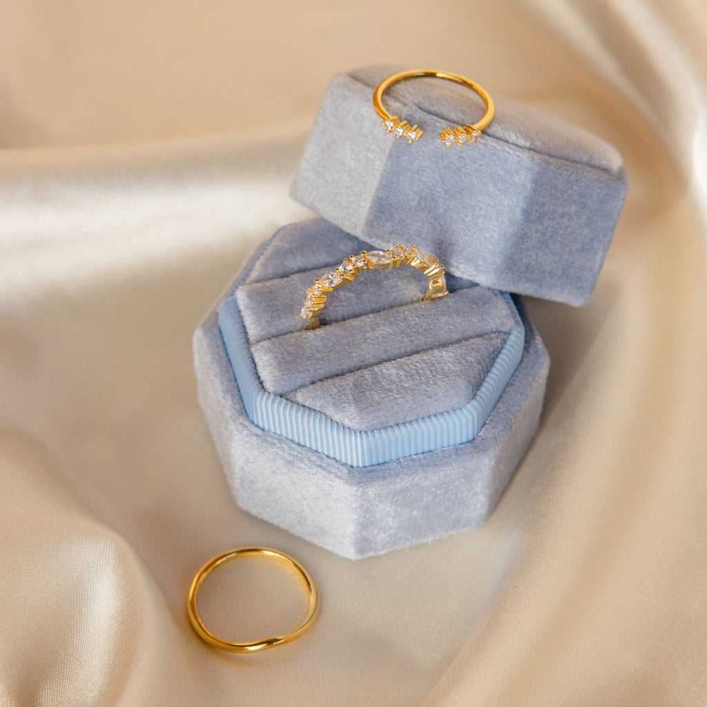 Two gold rings, one with small stones, featured in the Mystery Ring Box on a light blue velvet case over satin fabric.