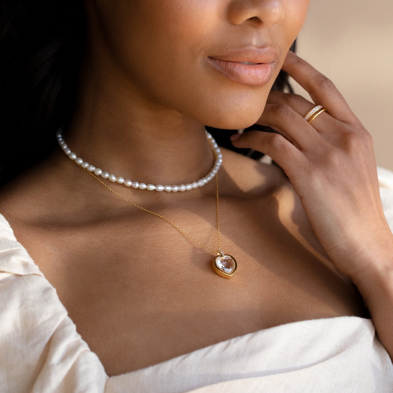 A woman in a cream top wears a pearl choker, a gold heart pendant, a delicate Crystal Locket Necklace, and a gold ring.