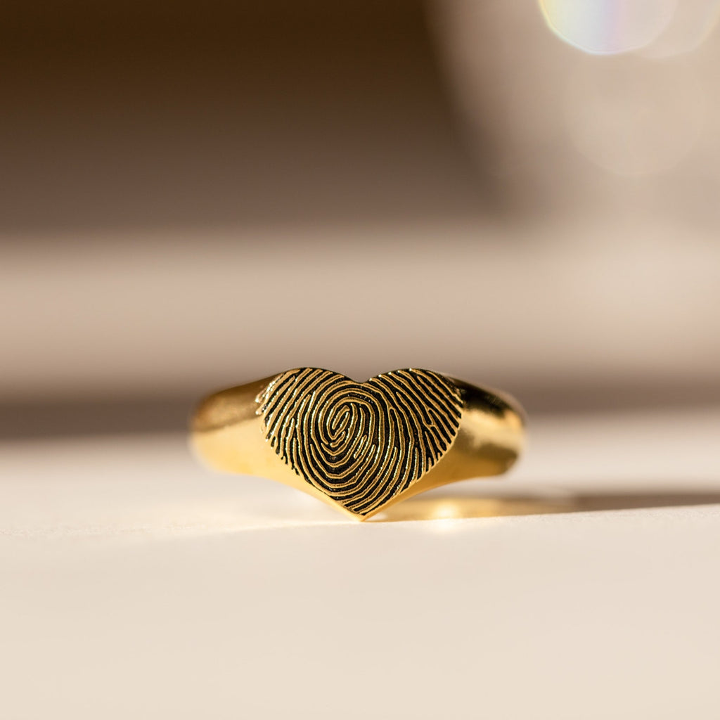 The Fingerprint Heart Signet Ring features a delicately engraved heart-shaped top with your unique fingerprint—a meaningful, personalized gift for mom. Displayed on a light surface, it’s the perfect keepsake.