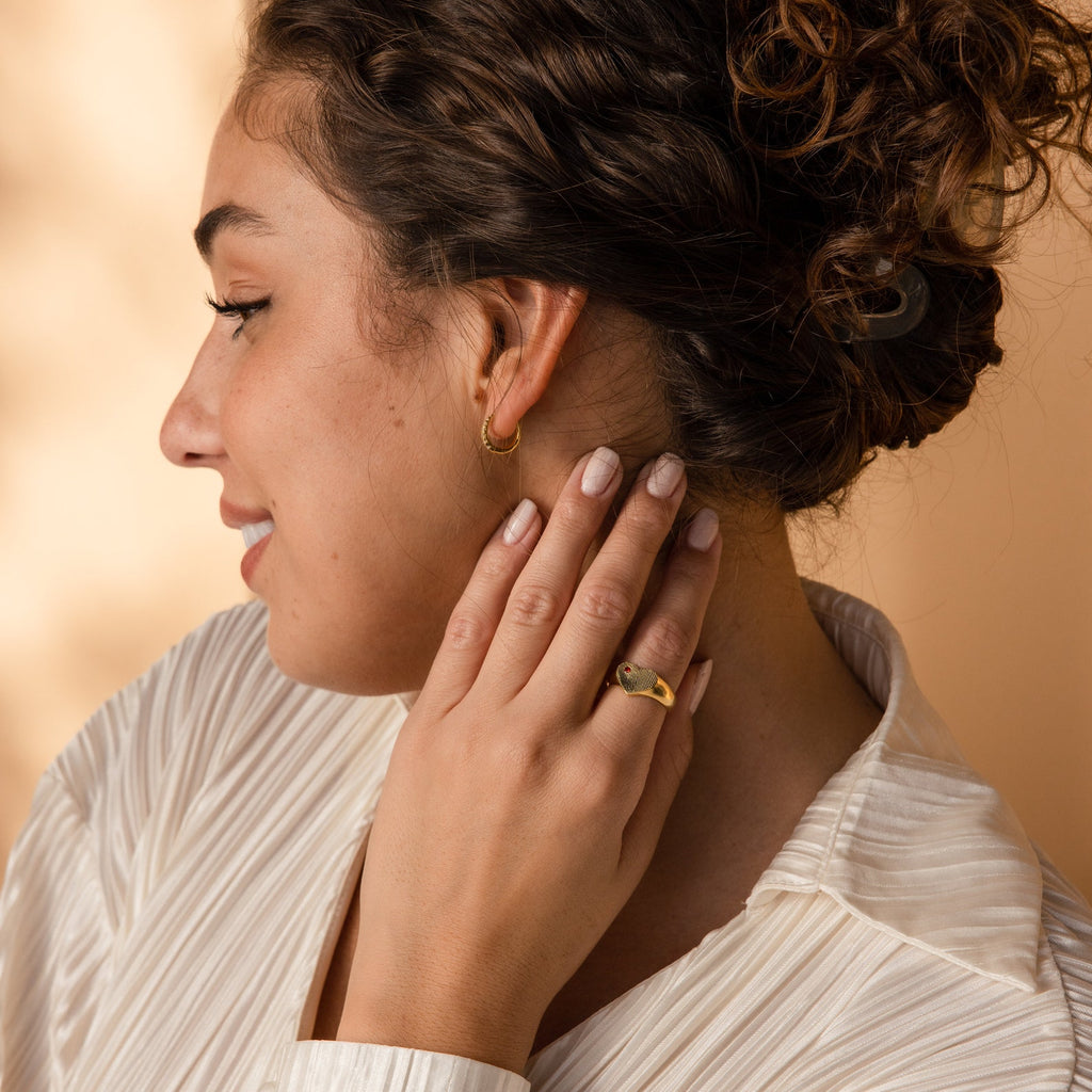Woman with curly hair in a white blouse touches her ear, showcasing gold earrings and the Fingerprint Heart Signet Ring—a unique and elegant piece that's a thoughtful gift for mom.