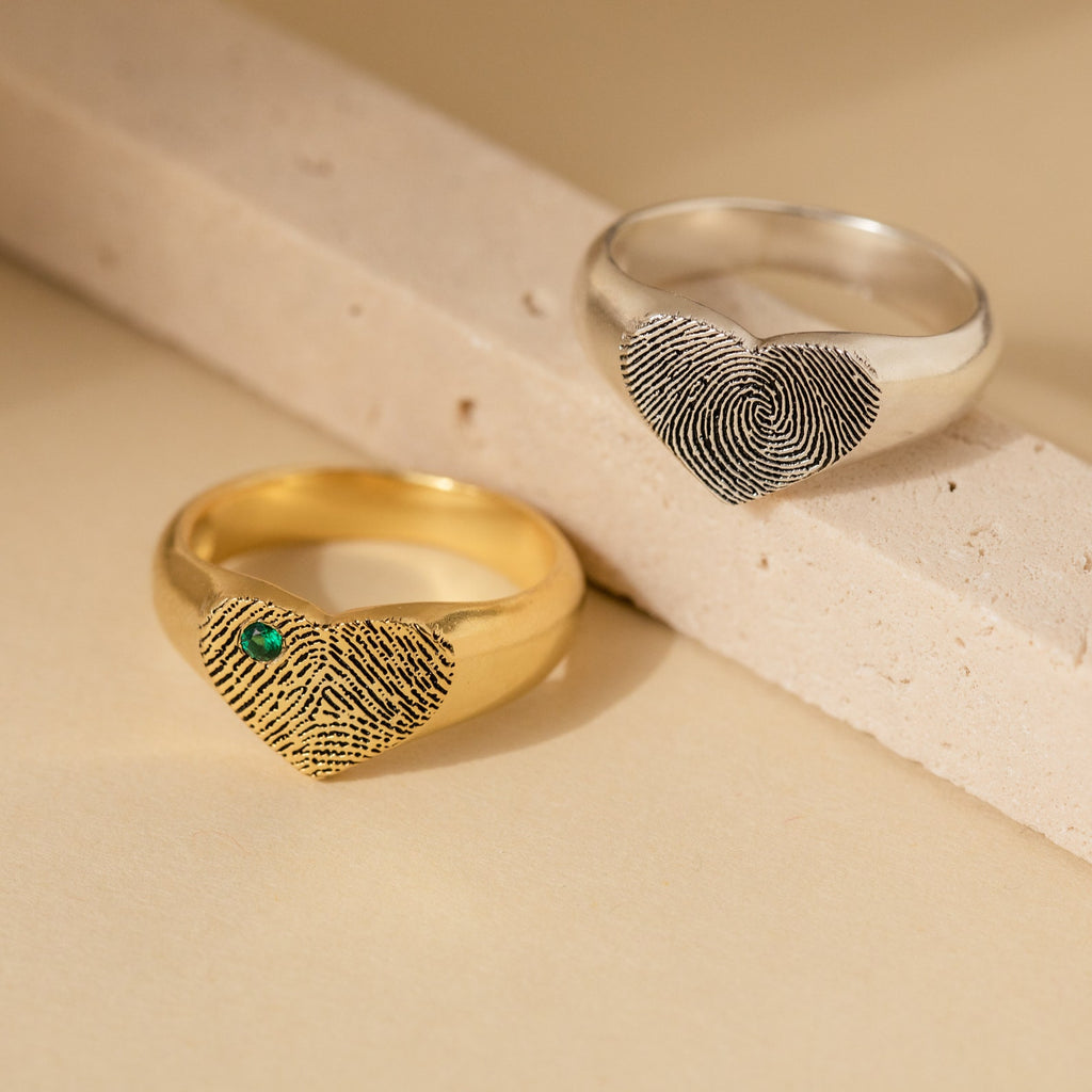 The Fingerprint Heart Signet Ring—one gold with a green stone, one silver—features personalized fingerprint designs on heart-shaped faces, resting on a beige surface. A unique, meaningful gift for mom.