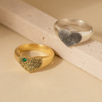 The Fingerprint Heart Signet Ring—one gold with a green stone, one silver—features personalized fingerprint designs on heart-shaped faces, resting on a beige surface. A unique, meaningful gift for mom.