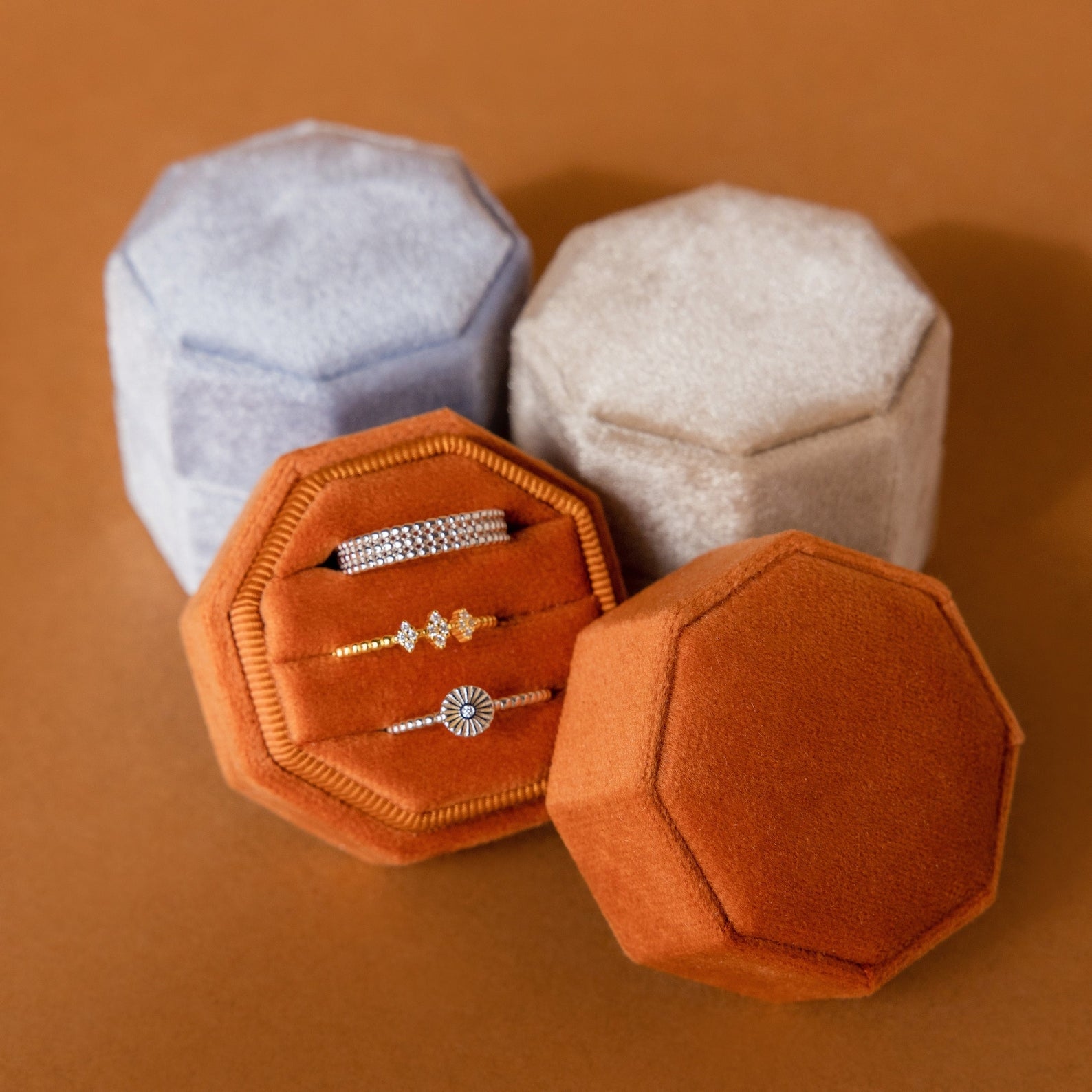 Three Mystery Ring Box cases in gray, beige, and orange velvet display gold and silver rings on a brown surface.
