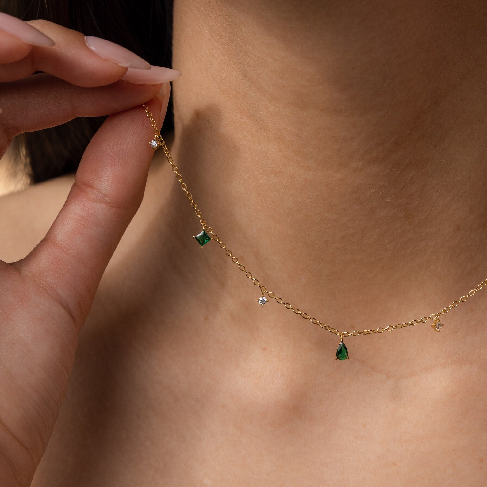 A hand holds the Larissa Emerald Station Necklace, a delicate, vintage-inspired piece with green and clear gemstone charms, gently resting on a person’s neck.