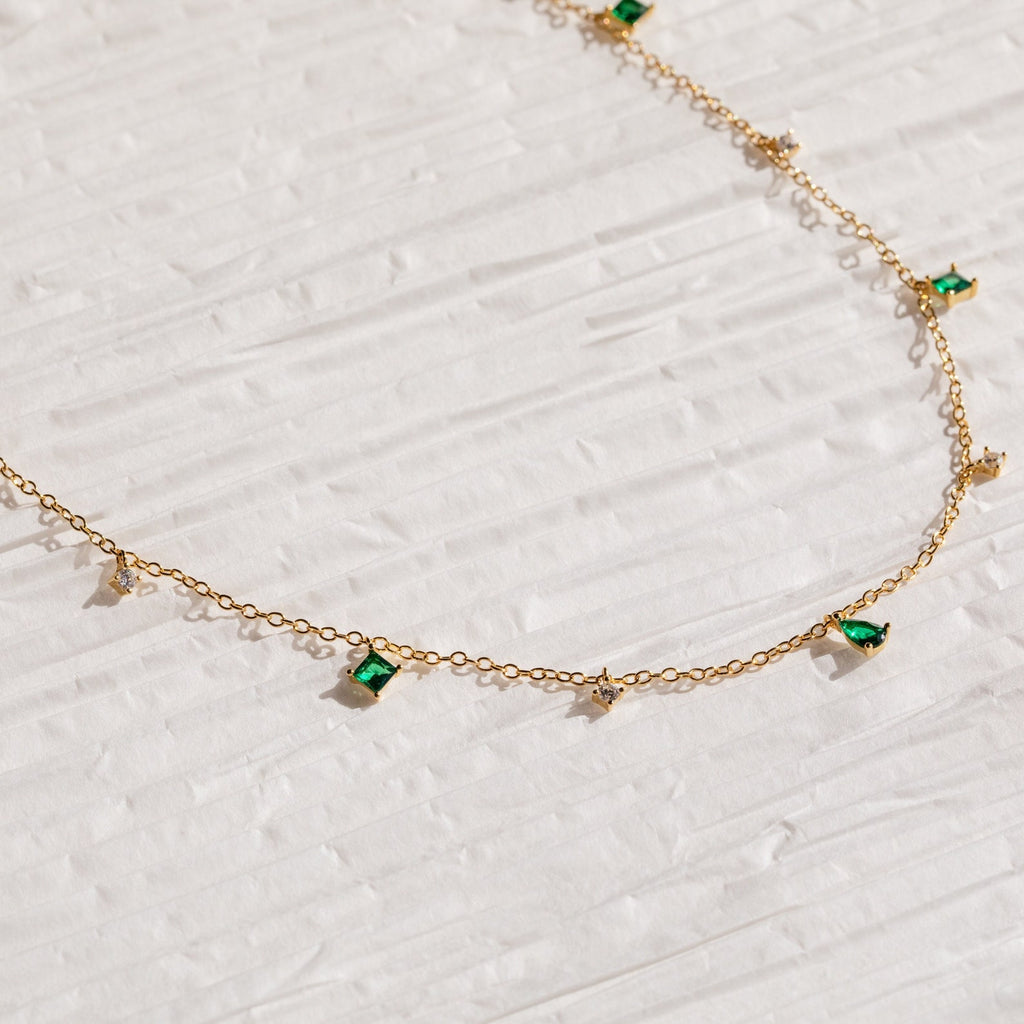 The Larissa Emerald Station Necklace features small green and clear gemstone charms, offering vintage-inspired elegance that shines on a textured white surface.