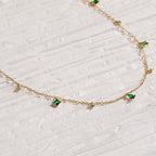 The Larissa Emerald Station Necklace features small green and clear gemstone charms, offering vintage-inspired elegance that shines on a textured white surface.