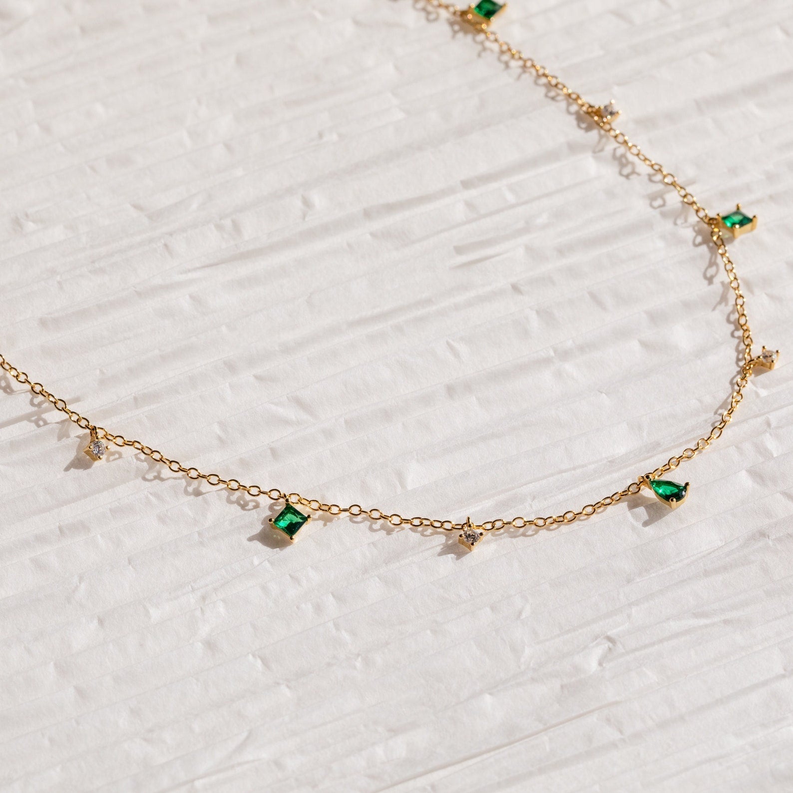 The Larissa Emerald Station Necklace features small green and clear gemstone charms, offering vintage-inspired elegance that shines on a textured white surface.