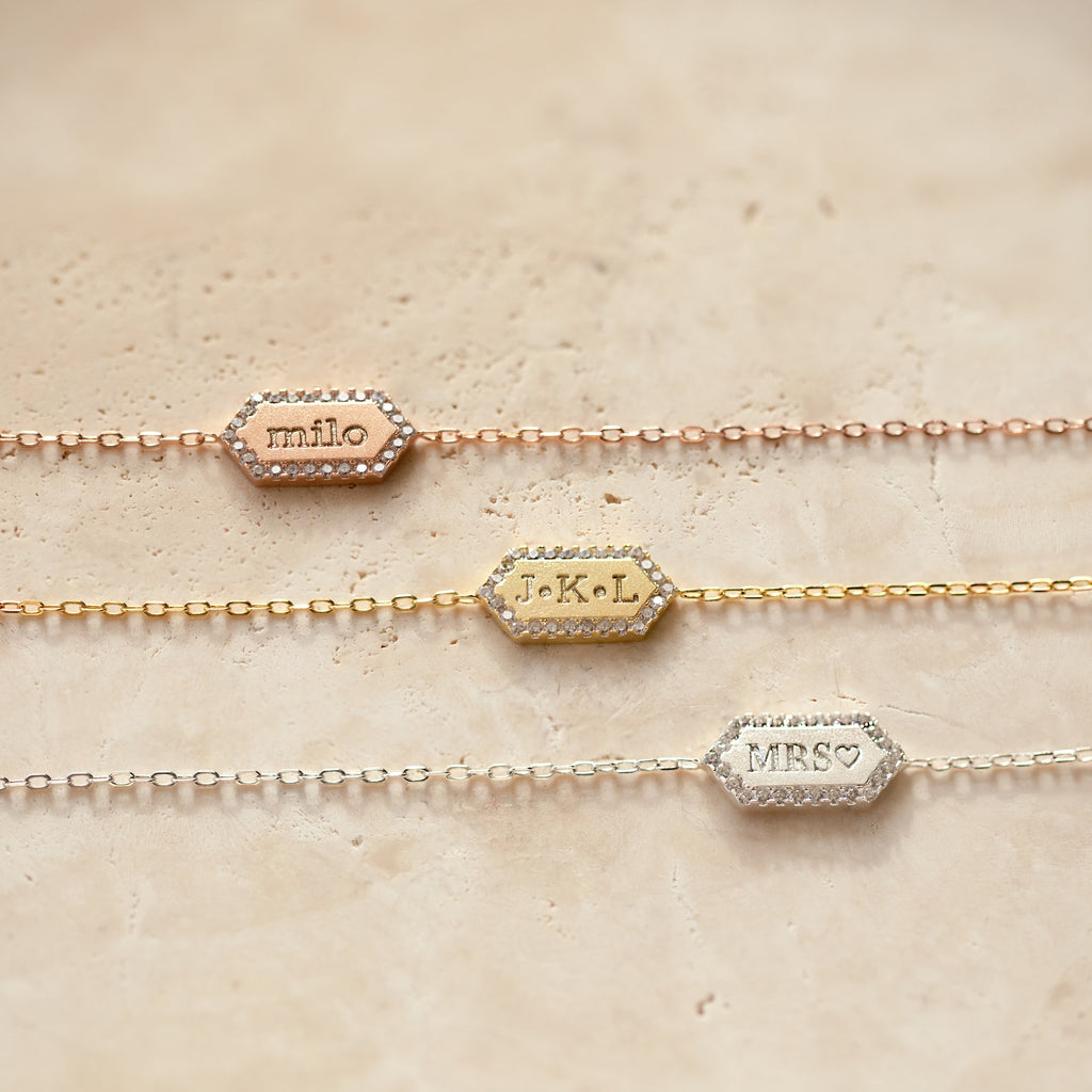 The Custom Pave Hexagon Bracelet, shown with “milo,” “J.K.L,” and “MRS♡” engravings on delicate chains, rests on beige. Personalize this thoughtful anniversary gift with your own custom engraving.