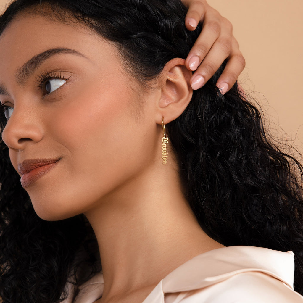 Woman with curly hair wears Old English Dangling Name Earrings reading "Brooklyn," as she touches her ear and gazes to the side.