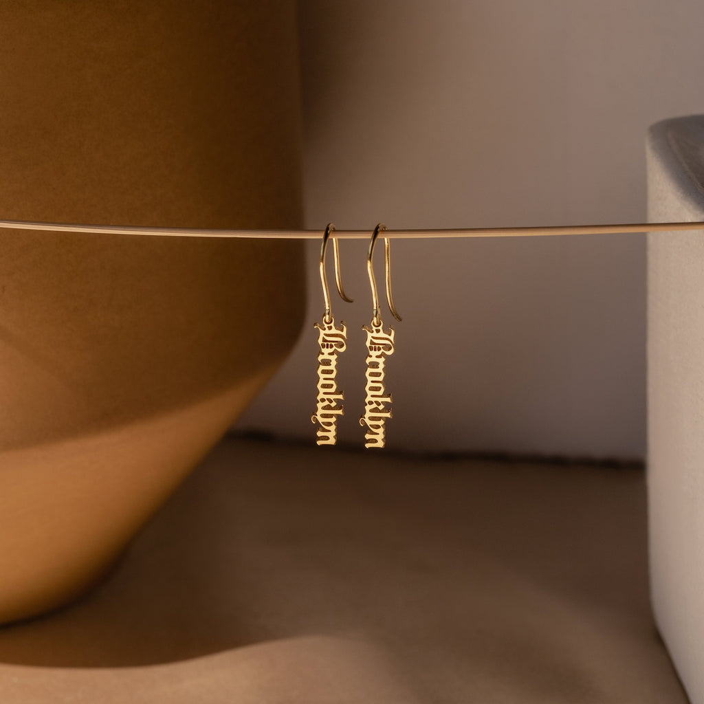 Old English Dangling Name Earrings feature your name in bold script, suspended from a horizontal rod against a neutral backdrop—perfect as handmade drop earrings or a unique personalized gift.