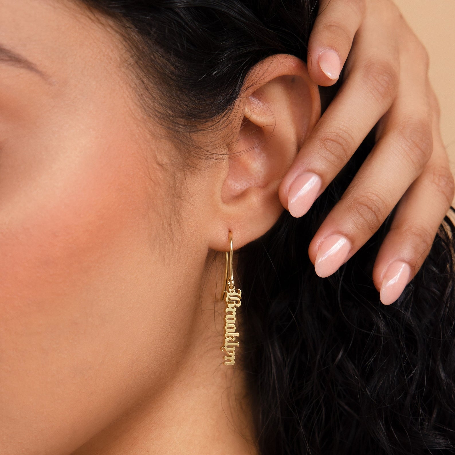 Close-up of a woman's ear wearing Old English Dangling Name Earrings that spell Brooklyn, with her hand near her ear—a perfect custom gift.