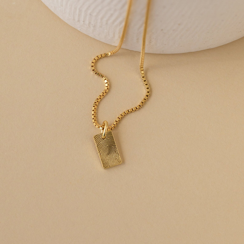 The Mini Fingerprint Tag Necklace features a rectangular, engraved pendant for a personalized touch—elegantly displayed on a beige surface. A thoughtful Mother's Day gift.