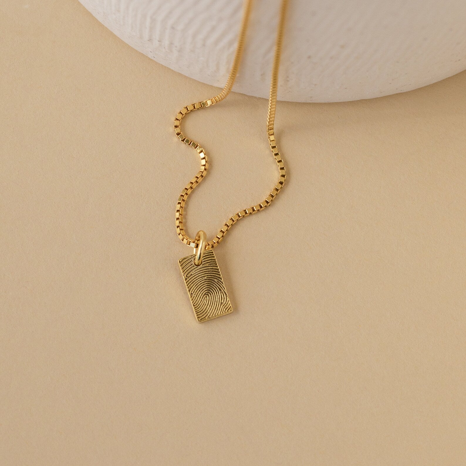 The Mini Fingerprint Tag Necklace features a rectangular, engraved pendant for a personalized touch—elegantly displayed on a beige surface. A thoughtful Mother's Day gift.