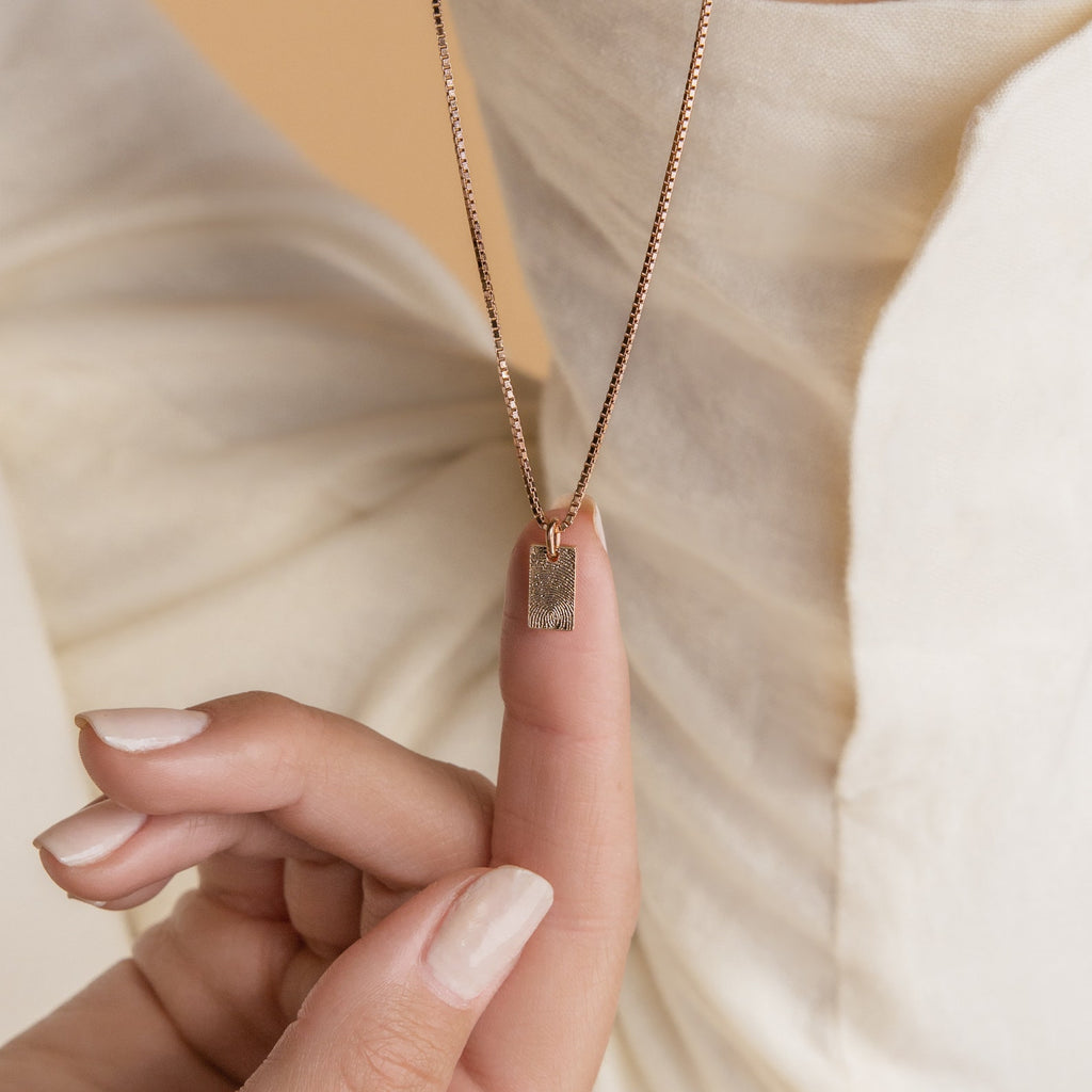 A hand displays the Mini Fingerprint Tag Necklace with a gold rectangular pendant against cream textured fabric—an elegant, personalized choice for Mother’s Day.