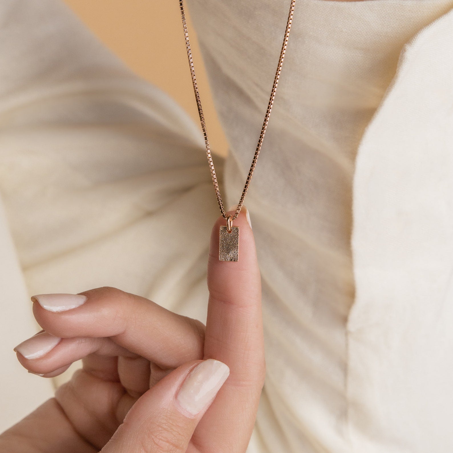 A hand displays the Mini Fingerprint Tag Necklace with a gold rectangular pendant against cream textured fabric—an elegant, personalized choice for Mother’s Day.