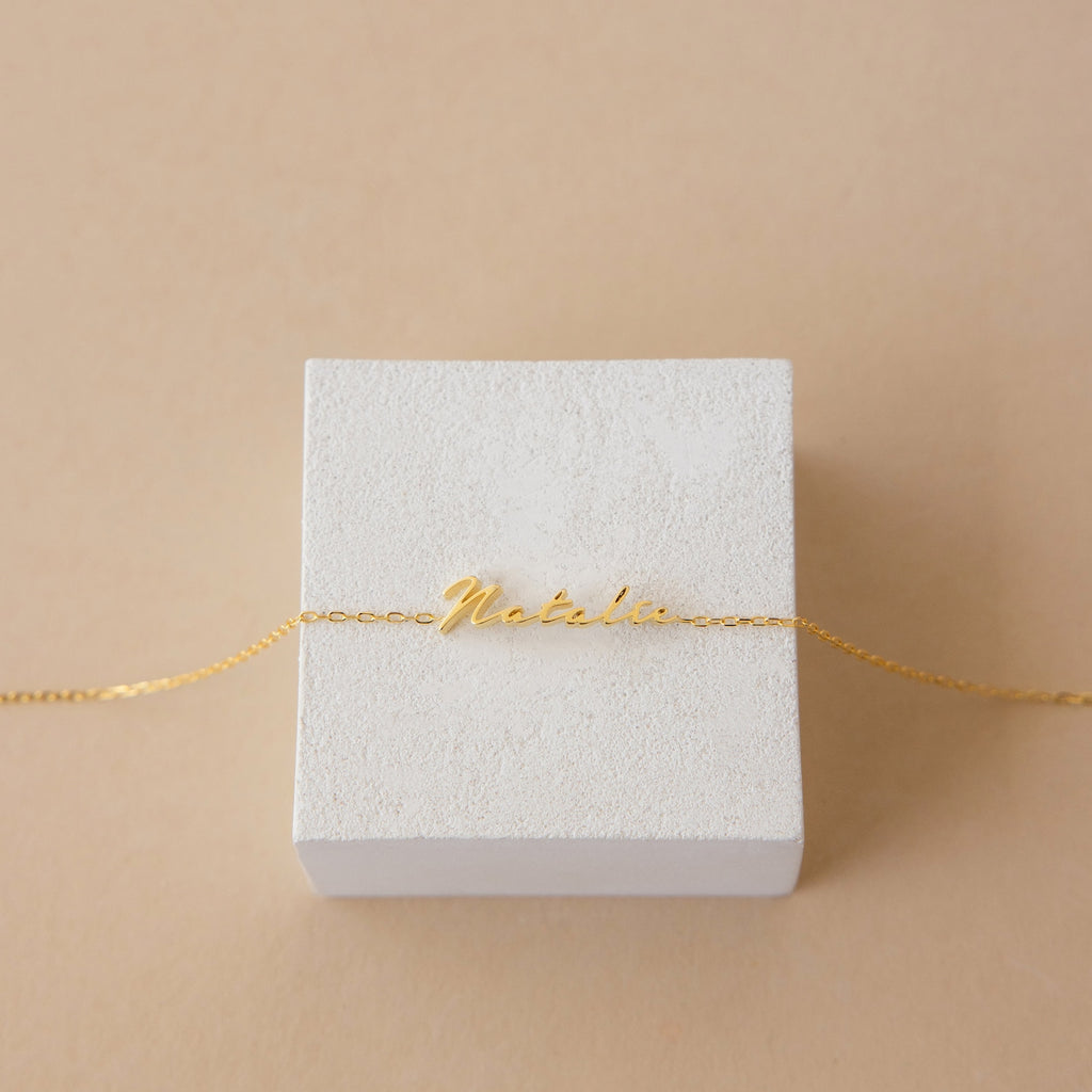 The Venice Tiny Name Bracelet, shown with "Natalia" on a white textured box and beige background, is a perfect piece of minimal jewelry.