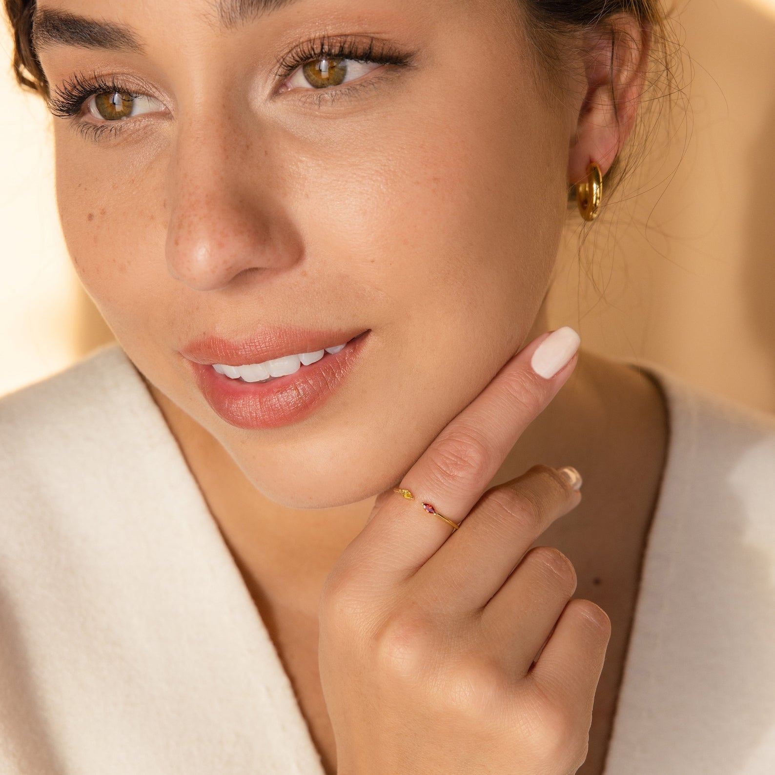 A woman with natural makeup wears gold hoop earrings and the Duo Marquise Birthstone Ring, smiling softly with her hand near her face.