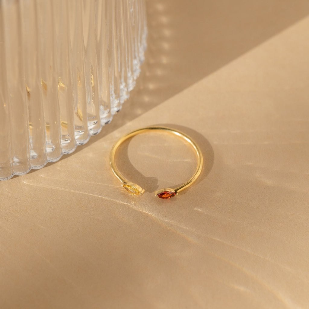 The Duo Marquise Birthstone Ring, featuring two small gemstones, rests on a beige surface beside a ribbed glass object.