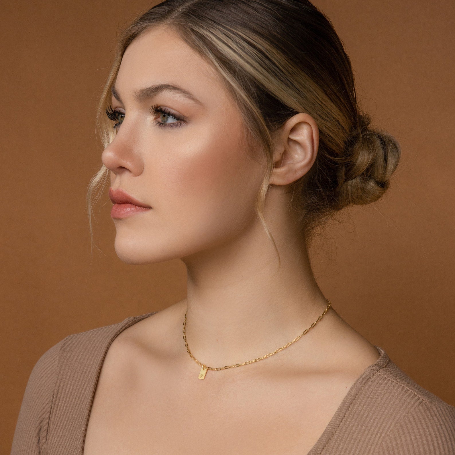 A blonde-haired woman with a low bun wears the Mini Initial Paperclip Necklace in gold, gazing left against a brown background.
