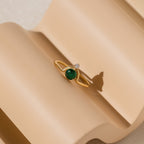 The Duo Vintage Birthstone Ring features a gold band with a green gemstone and a small clear stone, elegantly displayed on a beige, wavy surface—an ideal personalized Mother’s Day or new mom gift.