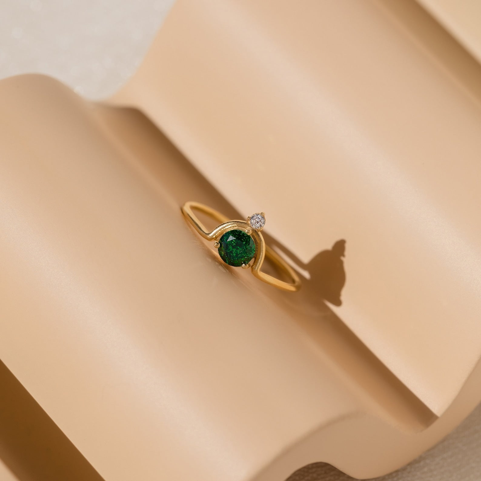 The Duo Vintage Birthstone Ring features a gold band with a green gemstone and a small clear stone, elegantly displayed on a beige, wavy surface—an ideal personalized Mother’s Day or new mom gift.