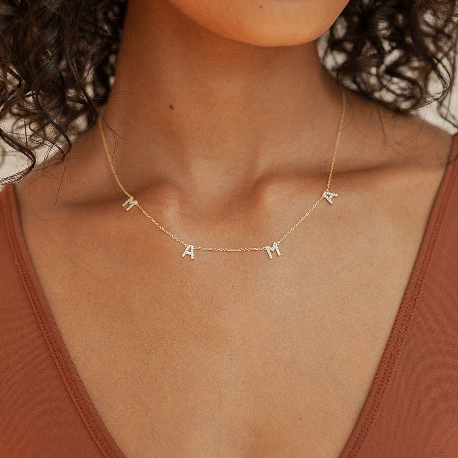 A person wearing a Mama Necklaces piece with letter charms spelling MAMA, paired with a brown V-neck top—an ideal 925 Sterling Silver gift for Mom.